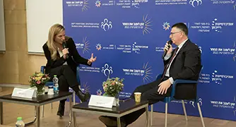 שני אנשים יושבים על כורסאות על במה. אחד מחזיק מיקרופון